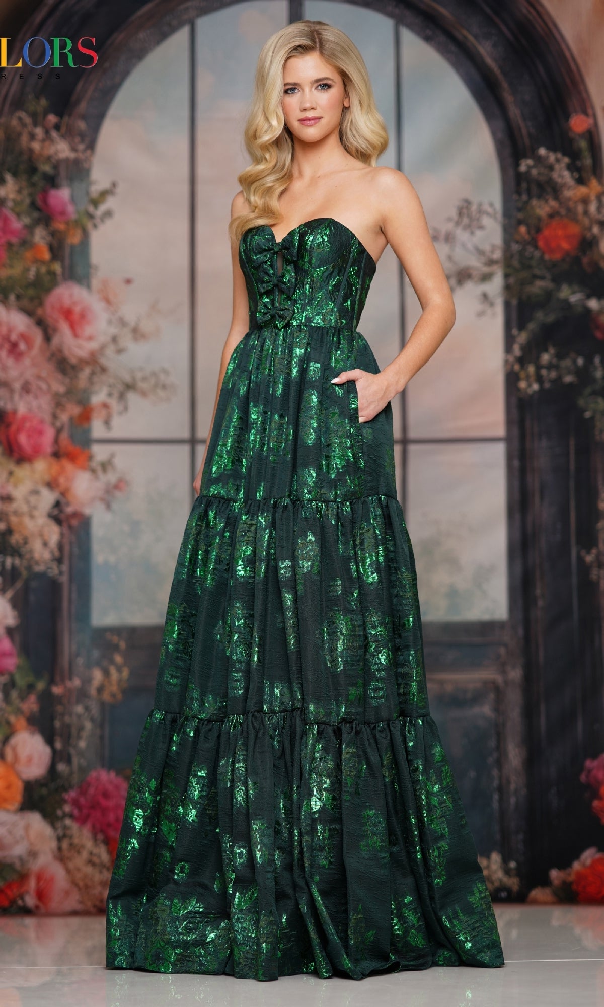 A woman wearing the Colors Dress 3794, a strapless long prom gown with a corset bodice and tiered flowy skirt, stands before a floral backdrop with arched windows.